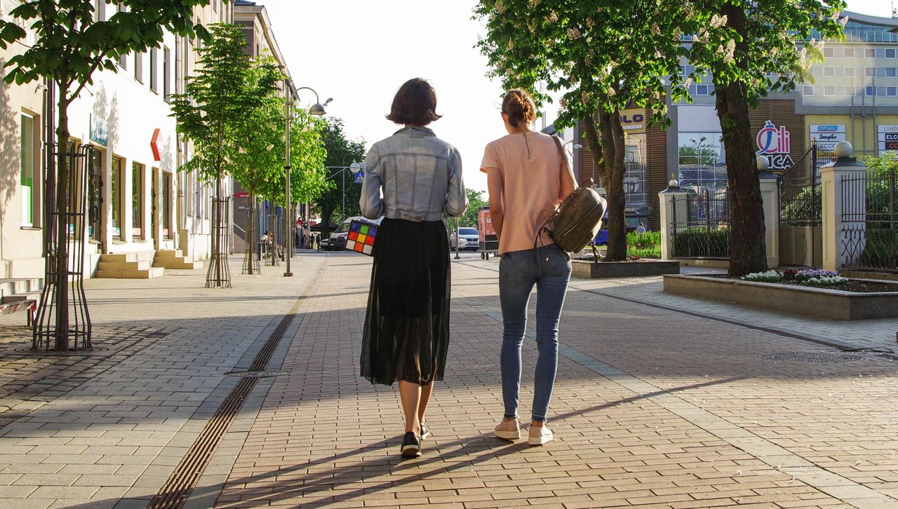 Two adult women walking together during a wellbeing support visit Two women enjoy a sunny walk down a lively urban street lined with buildings and trees.